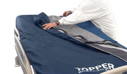 A person in a white uniform is adjusting a navy blue and gray medical monitoring mattress cover labeled 'TOPPER' on a hospital bed against a white background.