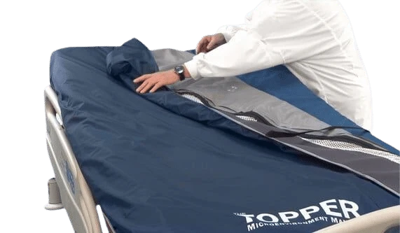 A person in a white uniform is adjusting a navy blue and gray medical monitoring mattress cover labeled 'TOPPER' on a hospital bed against a white background.