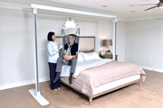 Person using a medical lift to assist another person onto a bed in a home setting.