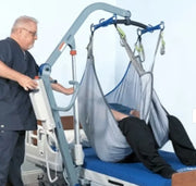 A medical transfer lift in use, showing a caregiver operating the gray and blue lift table while a person is being transferred in a light gray sling from a hospital bed.