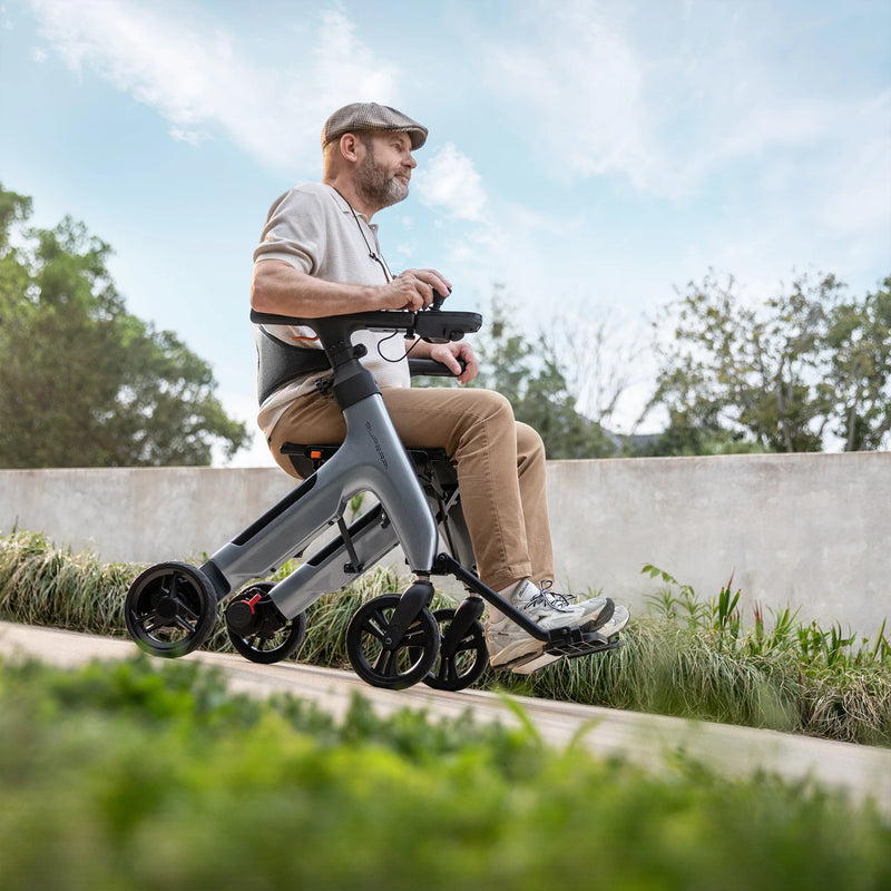 A man wearing a cap sits on a gray three-wheel electric wheelchair outdoors on a paved path with green grass and trees in the background.