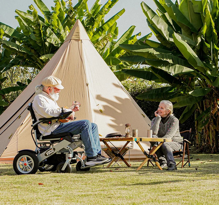Man in a wheelchair and woman sitting at a table in front of a tent with greenery around