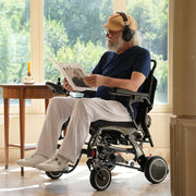 Man in a power wheelchair reading a magazine in a room with large windows.