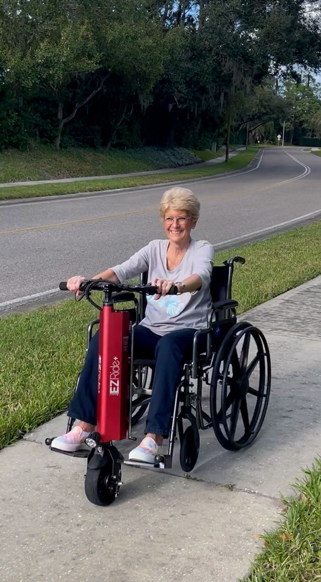 Woman using a wheelchair with a red scooter attachment on a sidewalk.