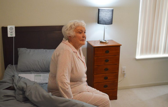Woman sitting on a bed in a bedroom with a wooden nightstand and lamp.