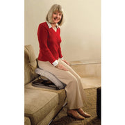An older woman sits on a beige upholstered sofa using a gray contoured lumbar support cushion with a black adjustable headrest. She is wearing a red knit sweater, a white collared shirt, and beige wide-leg trousers.
