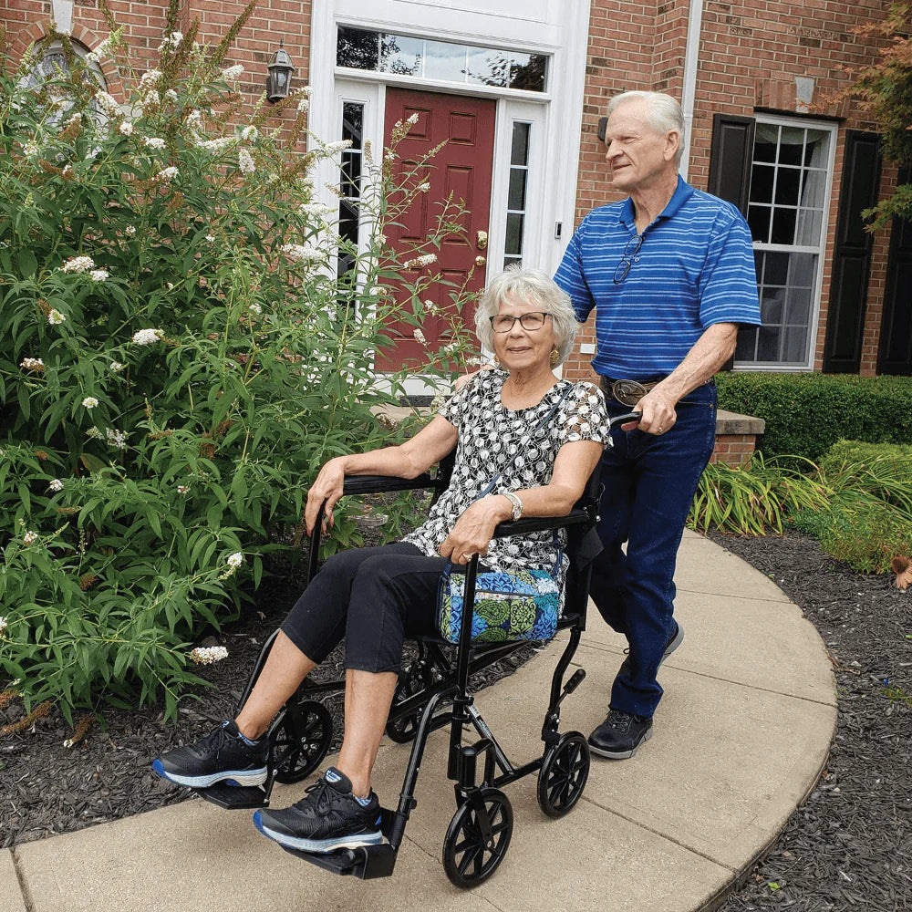 Man pushing a woman in a wheelchair on a sidewalk with a brick building and garden in the background