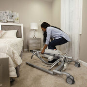 A woman in a bedroom sets up a hospital-style bed transport lift on a carpeted floor. The lift is gray with black wheels and white components, positioned near a bed, nightstand, lamp, and white curtain.
