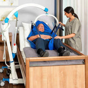 A hospital-style patient bed with a white and blue over-bed lift frame, showing a smiling elderly man reclining in a blue blanket while a nurse in gray scrubs adjusts the support straps beside the bed.