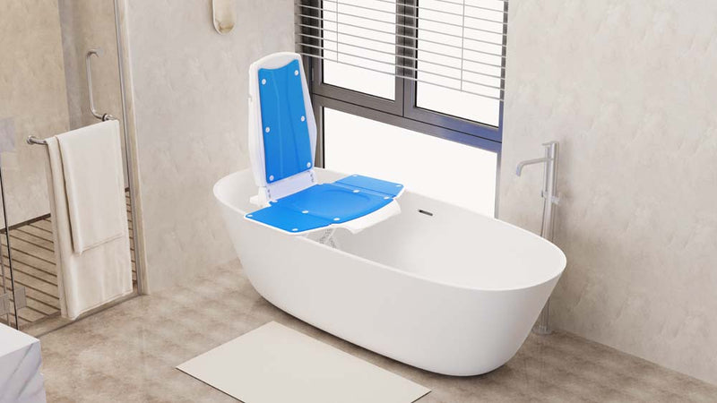 A white freestanding bathtub with a blue and white raised side seat attachment, shown in a modern beige bathroom with a window, shower glass, towel, and floor mat.