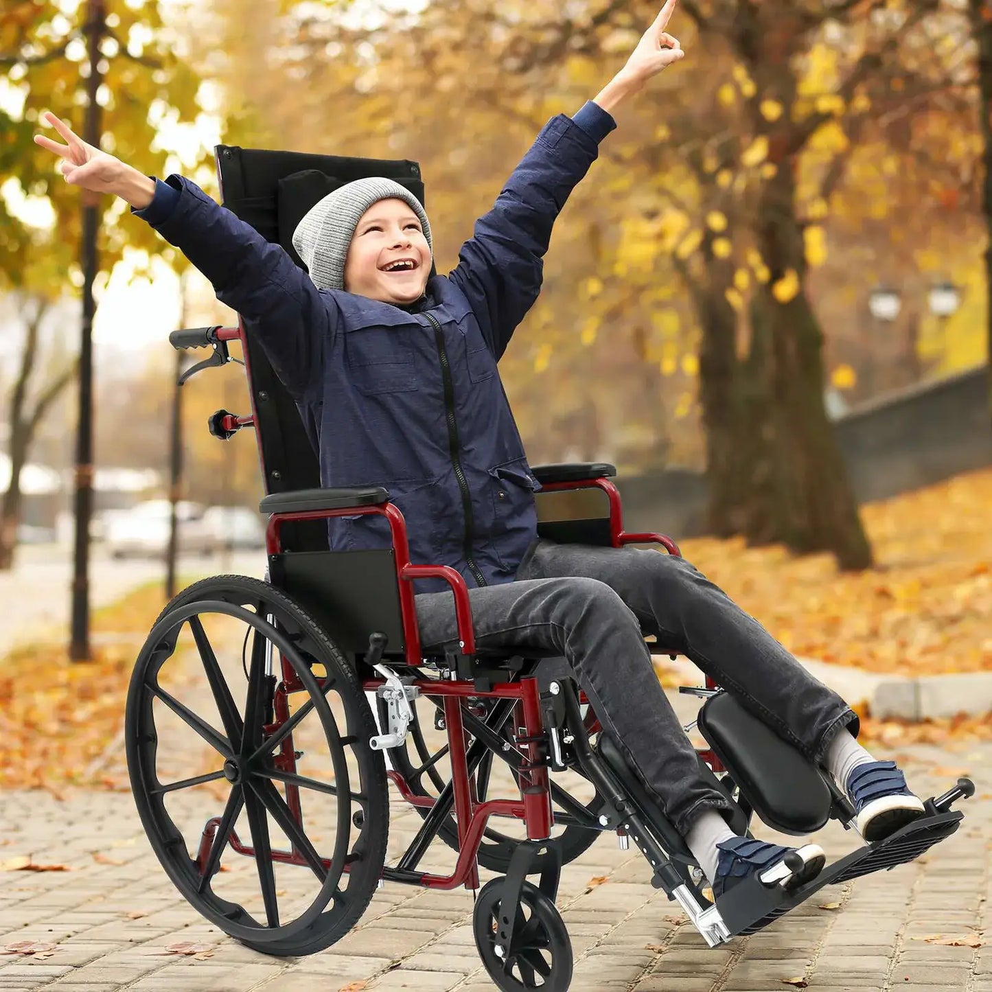 Child in a wheelchair with arms raised in an outdoor setting with trees and fallen leaves.