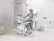 Woman using a shower chair in a bathroom with another person assisting her.