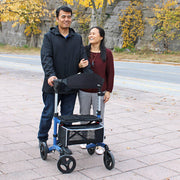 Man pushing a walker with a woman standing next to him on a sidewalk with autumn foliage.