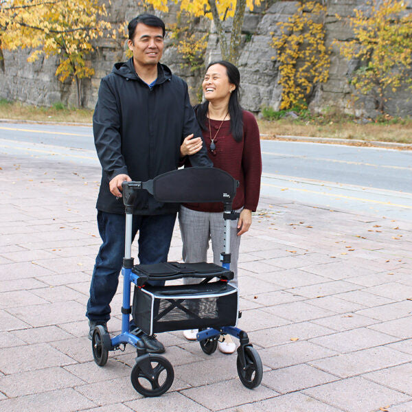 Man pushing a walker with a woman standing next to him on a sidewalk with autumn foliage.