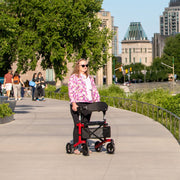 Person using a red and black rollator on a city sidewalk with buildings and trees in the background.