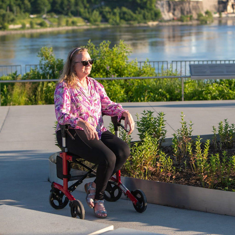 Person using a mobility scooter by a waterfront with greenery and a building in the background.