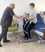 A person using a blue and gray sit-to-stand transfer table in a clinical room, assisted by a healthcare provider standing beside them.