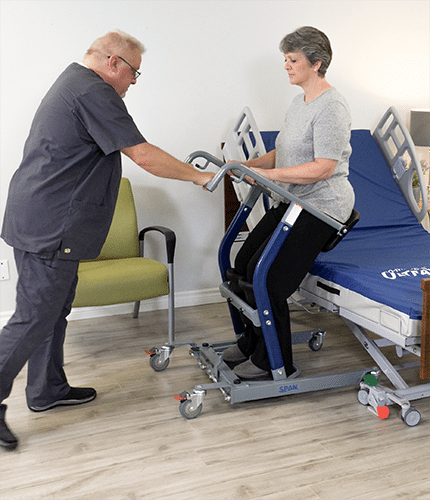 A person using a blue and gray sit-to-stand transfer table in a clinical room, assisted by a healthcare provider standing beside them.