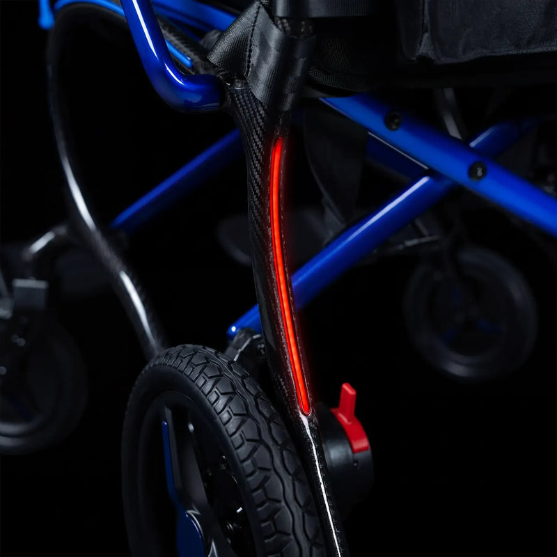 Close-up of a wheelchair frame with a bright red illuminated safety light mounted on a black carbon-fiber-style vertical bar, set against a dark background.