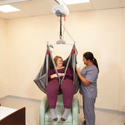 A woman sits in a suspended hospital-style lift chair while a nurse in gray scrubs stands beside it in a clinical room.