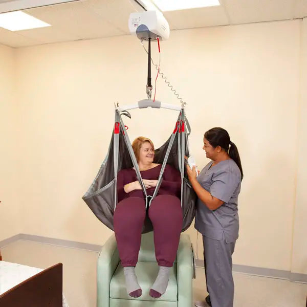 A woman sits in a suspended hospital-style lift chair while a nurse in gray scrubs stands beside it in a clinical room.