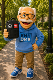 A person in a mascot costume stands on a tree-lined sidewalk holding a black first aid pouch. The costume includes a blue hoodie with 'ENH DME' printed on the front, tan pants, blue sneakers, and a white mustache.