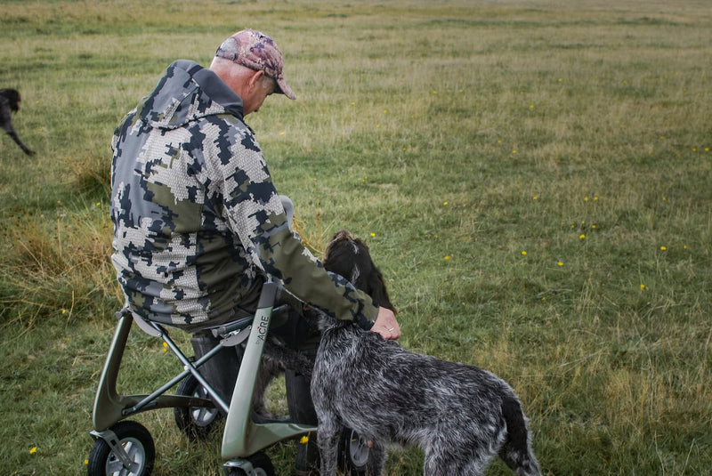 A man in a camouflage hunting jacket and cap sits on a rollator in an open grassy field, petting a black and white steelhead trout dog.