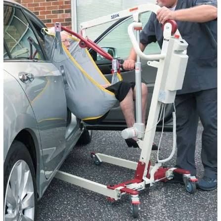 A motorized patient transfer lift being used to help a person get into a gray car. The lift has a white frame with red wheels and handles, and a gray transfer sling with yellow trim.