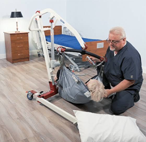 A person in dark scrubs kneels on a wood floor next to a hospital bed, handling a gray patient transfer bed with a white frame and red base. A pillow is in the foreground, and a bedside table with a lamp is visible in the background.