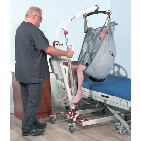 A healthcare worker assists a patient in a hospital bed using a motorized patient lift with a hanging mesh bed. The lift is white with red accents and is shown in a clinical room setting.