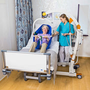 A geriatric patient in a blue hospital bed under a white and orange patient transfer frame, assisted by a healthcare worker in a bright room with wood floors and patterned curtains.