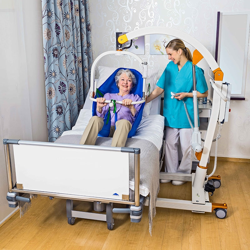 A geriatric patient in a blue hospital bed under a white and orange patient transfer frame, assisted by a healthcare worker in a bright room with wood floors and patterned curtains.