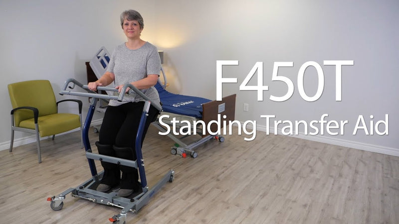 A woman using an F450T standing transfer aid in a bright room with wood floors, a green chair, and a treatment table. Large white text on the right reads 'F450T Standing Transfer Aid'.