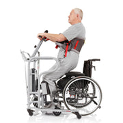 An older man in a gray outfit is shown seated in a motorized wheelchair transfer chair and wheelchair, photographed from the side on a clean white studio background. The transfer chair has a light gray frame with black and red harness components.