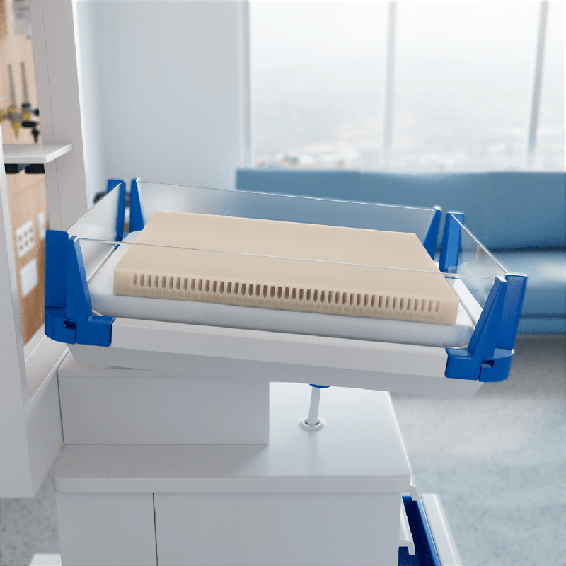 A beige mattress topper or cooling pad with a vented edge, enclosed in a clear protective cover and supported by blue and white mechanical arms, set in a modern room with a window and sofa in the background.