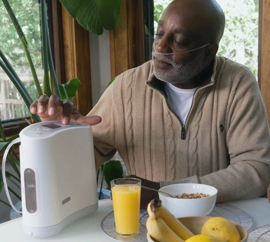 Man using a white appliance at a table with a glass of orange juice and bananas.