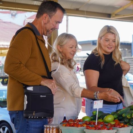 Three people shopping at an outdoor market with produce in the foreground.