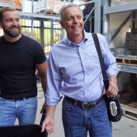 Two men standing in a warehouse setting, one holding a black bag.