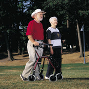 An older man wearing a white cowboy hat uses a four-wheel walker with a basket in a park, accompanied by an older woman in a black, white, and gray top.