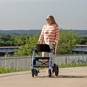 Woman walking with a walker outdoors on a path with greenery and a bridge in the background.