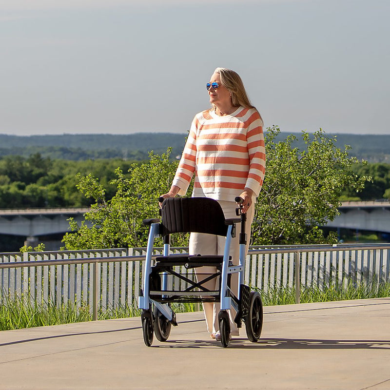 Woman walking with a walker outdoors on a path with greenery and a bridge in the background.