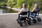 Two women using mobility scooters by a body of water with trees in the background.
