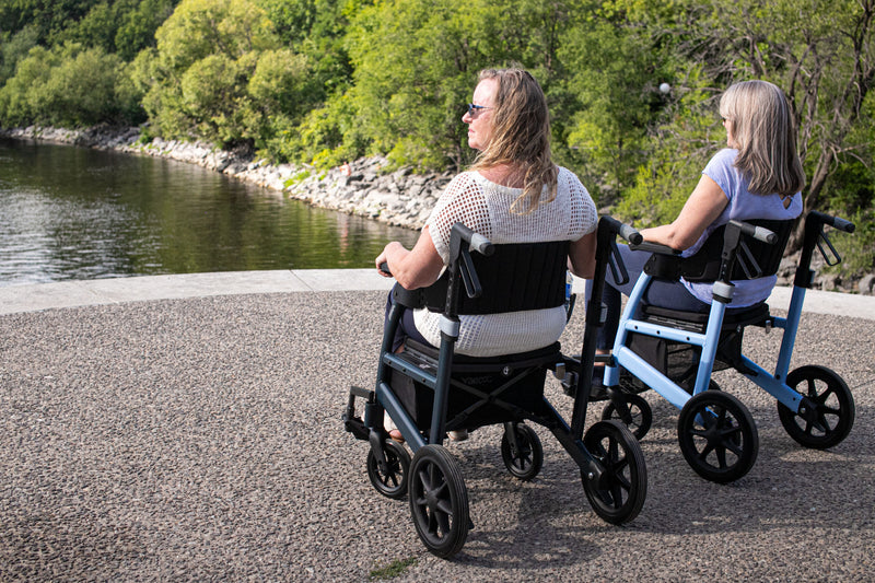 Two women using mobility scooters by a body of water with trees in the background.