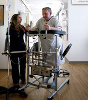 A patient in a hospital gown using a walker assisted by a nurse in a bright clinic room.