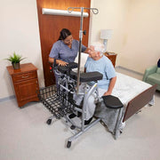 A caregiver assists an elderly man using a walker and transfer frame in a bedroom setting.