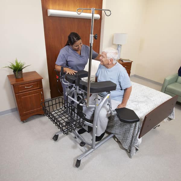 A caregiver assists an elderly man using a walker and transfer frame in a bedroom setting.