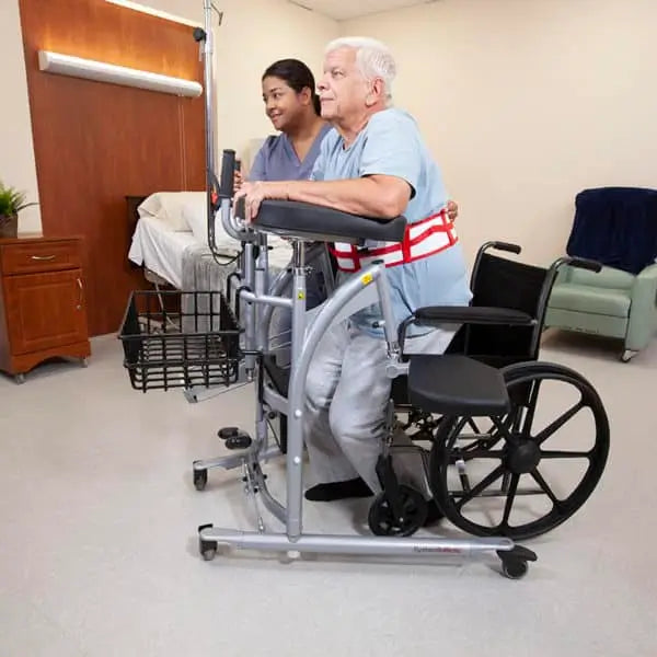 Person using a walker in a medical setting with another person assisting.